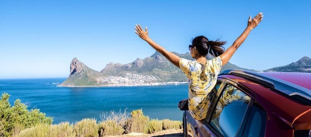 A woman enjoying a road trip on Chapman's Peak Drive in Cape Town, with her hands up, gazes out over the ocean from the car window