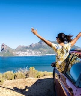 A woman enjoying a road trip on Chapman's Peak Drive in Cape Town, with her hands up, gazes out over the ocean from the car window