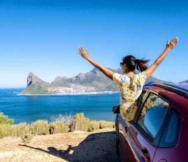 A woman enjoying a road trip on Chapman's Peak Drive in Cape Town, with her hands up, gazes out over the ocean from the car window